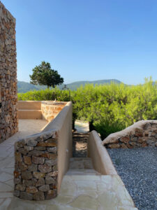Exterior view of a stunning 3-bedroom villa with 180-degree panoramic views of Cala San Vicente valley, showcasing its natural stone cladding, lush greenery, and peaceful hillside setting.
