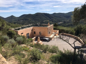 Exterior view of a stunning 3-bedroom villa with 180-degree panoramic views of Cala San Vicente valley, showcasing its natural stone cladding, lush greenery, and peaceful hillside setting.